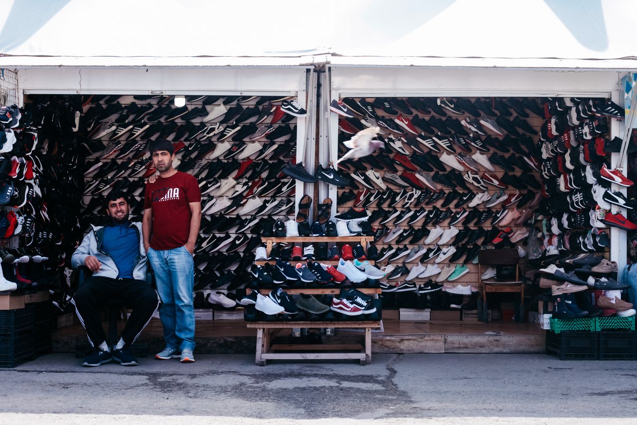 Colorful shoe stall at a street market with two men standing and seated. Wide assortment of footwear on display.